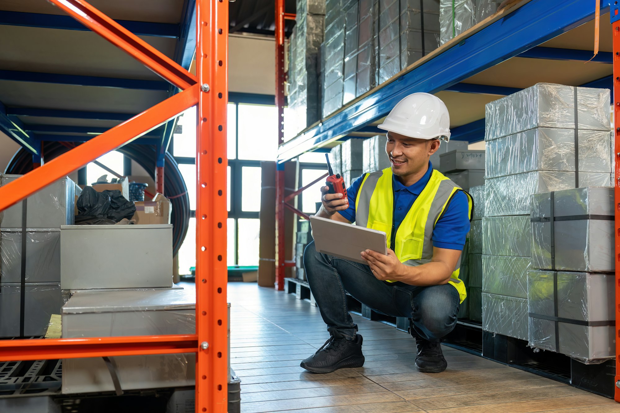 Asian male dispatcher checking packages while using walkie-talkie in distribution warehouse.