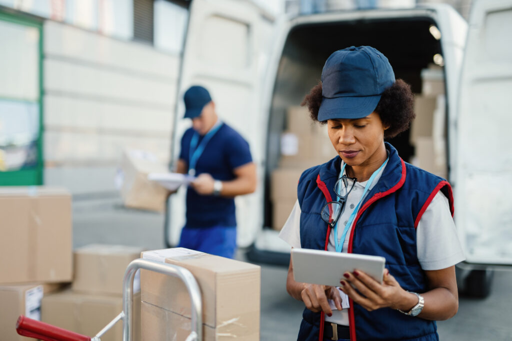 African American female worker making inventory of packages and organizing a delivery schedule on a digital tablet.  Her colleague is working in the background.